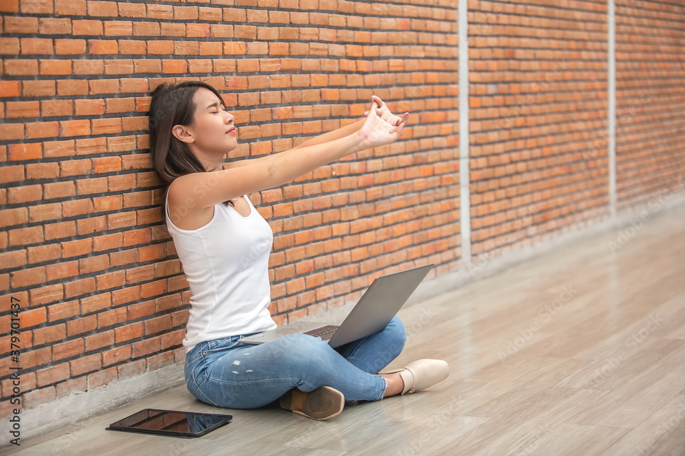 Beautiful young Asian woman sitting on floor and stretching arm up when working on laptop againts old brick wall
