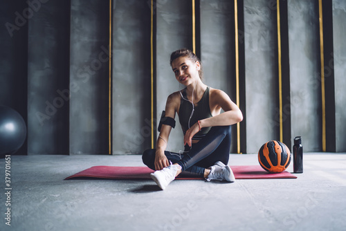 Half length portrait of professional prosperous woman in earphones resting after workout in gym, positive female athlete with ball looking at camera enjoying favorite songs from mobile phone