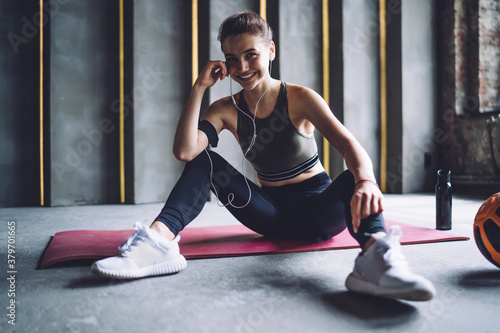 Half length portrait of pretty caucasian sportswoman satisfied with training results resting while listening music in earphones, happy young girl with ball using smartphone