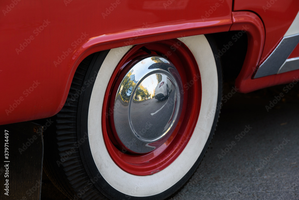 Closeup photo of the rear wheel of a magnificent retro car. The wheel has a red stamped disc in the body color, a large chrome hood and white wall.