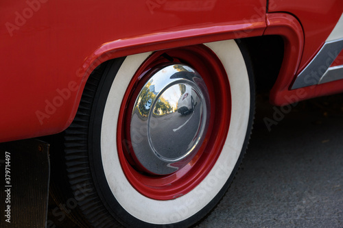 Closeup photo of the rear wheel of a magnificent retro car. The wheel has a red stamped disc in the body color, a large chrome hood and white wall. © Дмитрий Березнев