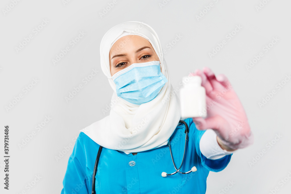 friendly Muslim doctor or nurse in a hijab, mask, gloves offering a pill to the patient on a gray background friendly Muslim doctor or nurse in a hijab, mask, gloves offering a pill to the patient on a gray background