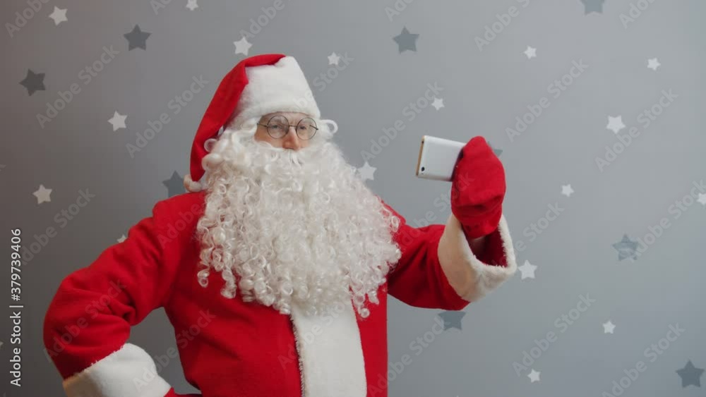 Man in Santa costume is holding smartphone touching screen taking selfie on grey background. Holidays, modern technology and people concept. Man in Santa costume is holding smartphone touching screen taking selfie on grey background. Holidays, modern technology and people concept.