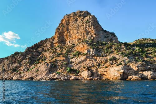red orange stone rock, sharp mountain with green grass and tress stands on shore of Black sea on a bright sunny day, summer sky with clouds