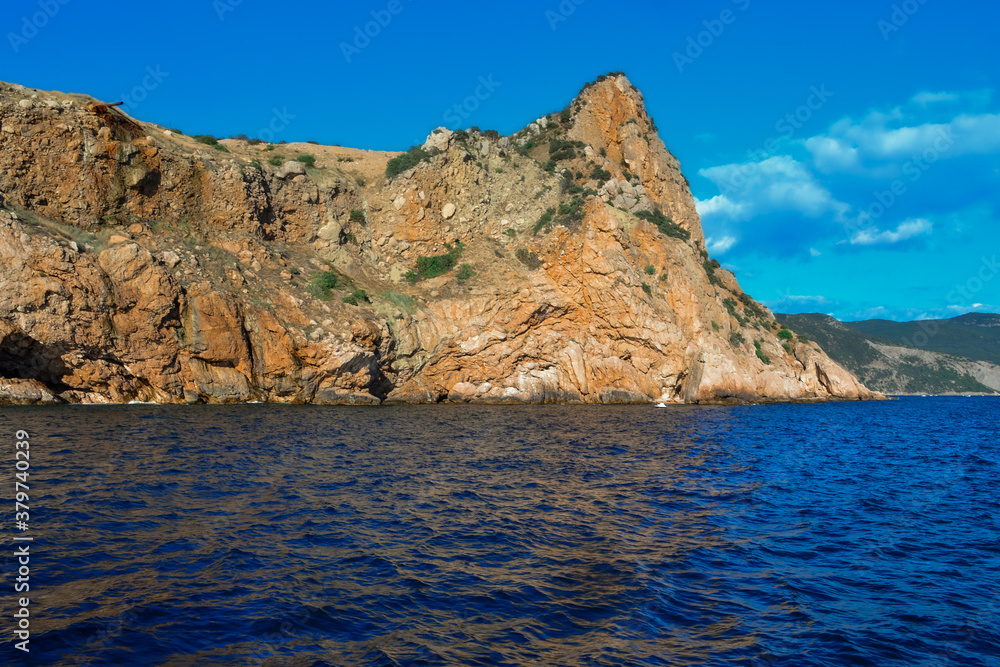 red stone rock stands on shore of the blue sea on a bright sunny day, summer sky with clouds