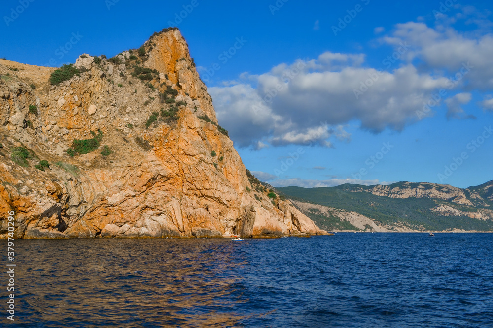 red orange stone rock, sharp mountain stands on shore of Black sea on a bright sunny day, summer sky with clouds