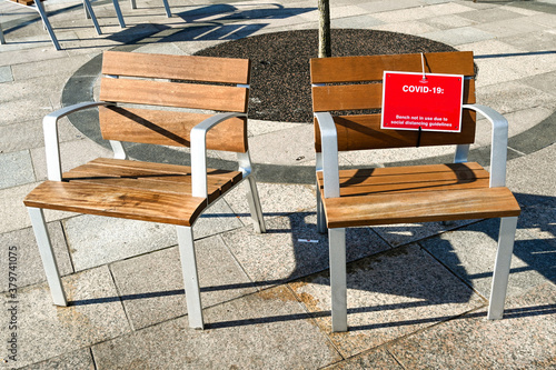 Two seats outdoors on a paved area. One has a sign informating people it is not in use due to social distancing measures for the Covid-19 pandemic. No people. photo