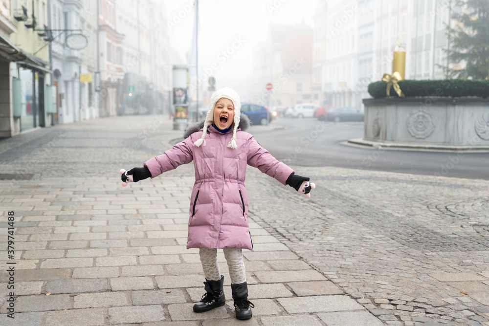 Cute liitle sad lost caucasian girl in warm long pink jacket and hat shouting loud on empty street of old european city Wels. Screaming angry alone scared child in town in Morning fog on backround