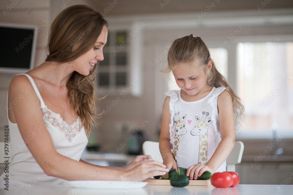 Naklejka premium Happy family mother and child posing at home. Beautiful young mom and little daughter having fun and preparing vegetables for salad in a white kitchen in a Scandinavian style interior. Healthy food.