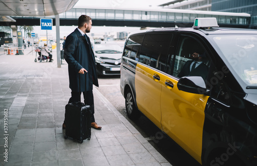 Serious man in elegant formal wear waiting for taxi cab near airport road getting to important business meeting with company partners, male commuter with suitcase bag choosing transfer auto