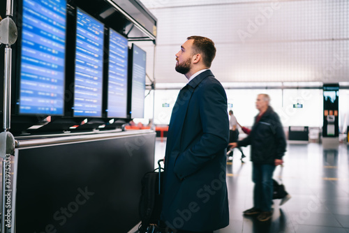 Caucasian male boss checking departure time and gate information on airport timetable standing near mockup stand with copy space area for advertising, businessman with luggage search flight schedule