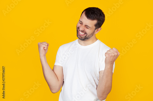 A young man with a beard in a white t-shirt is very happy and excited, making a winner gesture with raised hands, smiling and shouting success Celebration concept. Stands on isolated yellow background