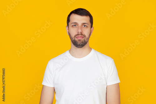 Young man with a beard in a white t-shirt with a serious expression on his face. Simple and natural looking into the camera. Stands on isolated yellow background