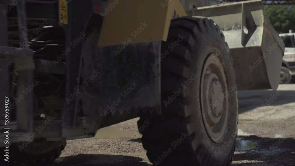 Large industrial tractor passing camera with loaded bucket. Loader with crushed stone riding outdoors on manufacturing site. Factory machinery working in sunlight. Large industrial tractor passing camera with loaded bucket. Loader with crushed stone riding outdoors on manufacturing site. Factory machinery working in sunlight.