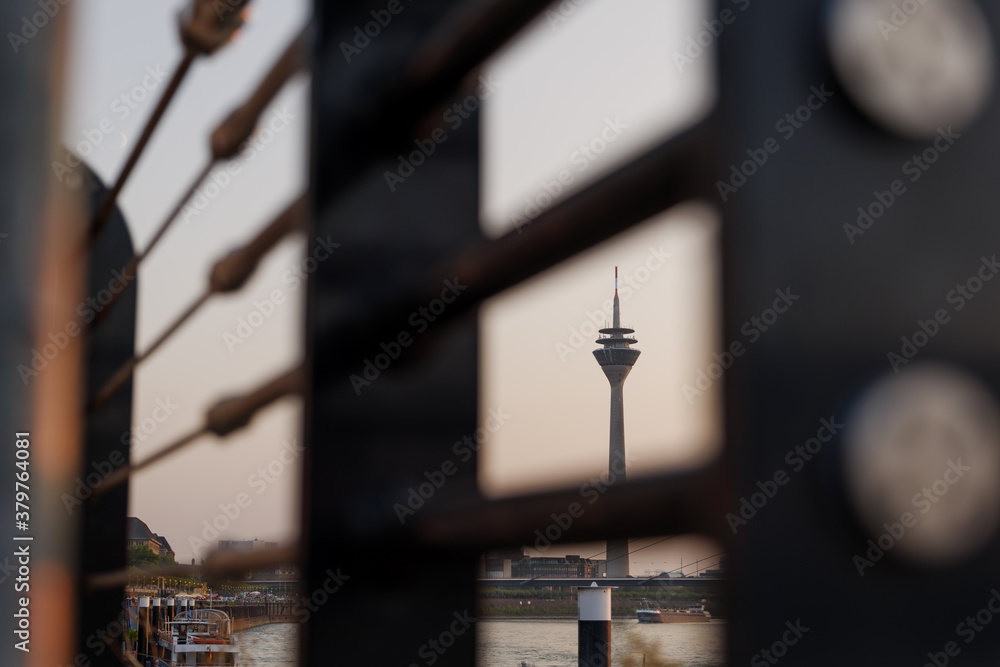 Selective focus view at Rheinturm, Rhine Tower, through pipe or wire cable of railing on promenade riverside of Rhein River in Düsseldorf, Germany during sunset with twilight sky. Selective focus view at Rheinturm, Rhine Tower, through pipe or wire cable of railing on promenade riverside of Rhein River in Düsseldorf, Germany during sunset with twilight sky.