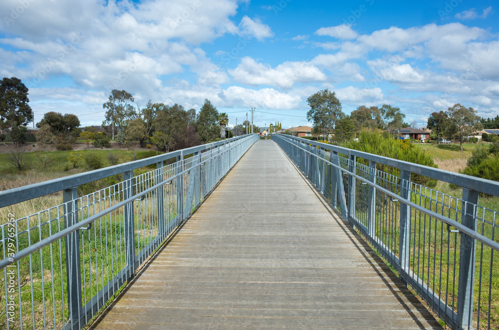A pedestrian footbridge/boardwalk over wetlands leads to an Australian neighbourhood with some residential houses in the distance. Skeleton Waterholes Creek, Melbourne, VIC Australia.