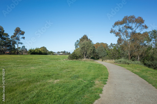 A concrete footpath/walkway in a large park with vast lawn areas, Australian native Eucalyptus trees and some houses in the distance. Werribee River Trail, Melbourne VIC Australia.