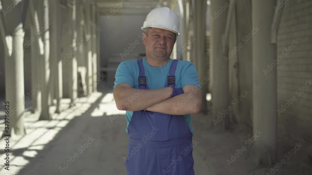 Confident positive man in uniform and helmet crossing hands and smiling at camera. Portrait of Caucasian builder posing on construction site in sunrays on sunny summer day. Confident positive man in uniform and helmet crossing hands and smiling at camera. Portrait of Caucasian builder posing on construction site in sunrays on sunny summer day.