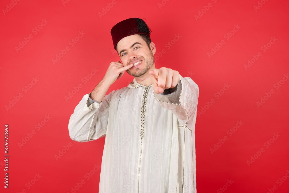 young caucasian muslim man wearing djellaba and traditional hat over red wall smiling cheerfully and pointing to camera while making a call you later gesture, talking on phone 