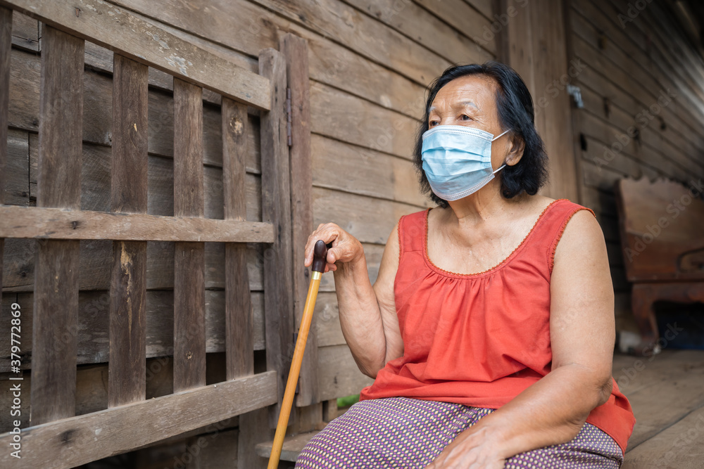 Thai elderly woman in round-necked sleeveless collar wearing medical mask for protect corona virus (covid-19) pandemic in wooden home