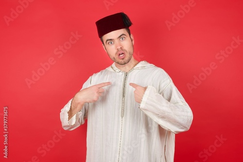 young caucasian muslim man wearing djellaba and traditional hat  indicates at hisself with puzzled expression, being shocked to be chosen to participate in competition, hesitates about something  photo