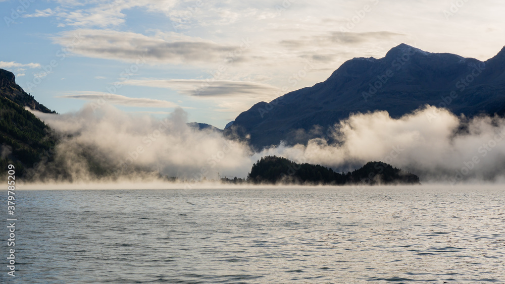Maloja mountain Pass, Switzerland. Landscape of the lake Sils in the morning during the fall time. The fog covers the lake and the shores. Traditional Swiss contest. Natural landscape Maloja mountain Pass, Switzerland. Landscape of the lake Sils in the morning during the fall time. The fog covers the lake and the shores. Traditional Swiss contest. Natural landscape