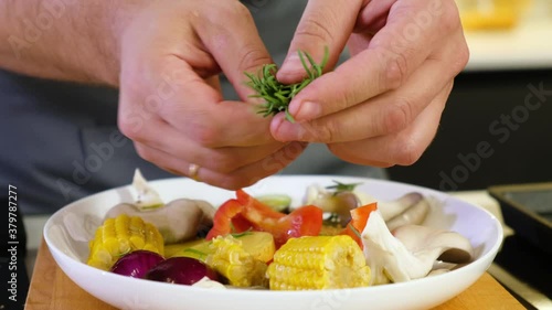 professional chef in kitchen prepares salad of peppers tomatoes onions mushrooms corn spices. man plucks leaves from sprig of rosemary with hands and sprinkles spices on the dish. Diet vegetable salad