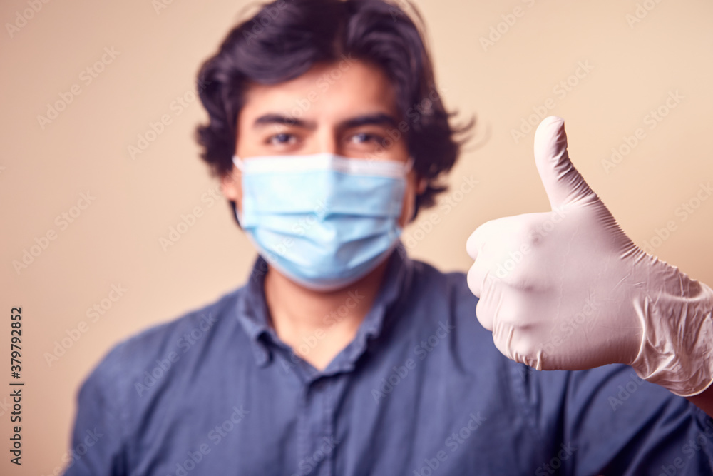 Un hombre joven sonrisa mientras usa mascarilla quirúrgica en el rostro y guantes levantando el pulgar. Un hombre joven sonrisa mientras usa mascarilla quirúrgica en el rostro y guantes levantando el pulgar.