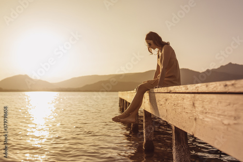 Pensive single Caucasian pretty young woman sitting on a pier of a lake looking down to the water at sunset - Vintage orange color mood