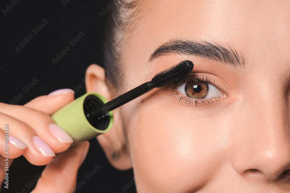 Beautiful young woman applying mascara against dark background, closeup