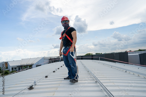strong look man wear sunglasses red hardhat black shirt blue jeans sneaker and hardness safety belt holding wrench working on metal sheet rooftop in beautiful day stock photo