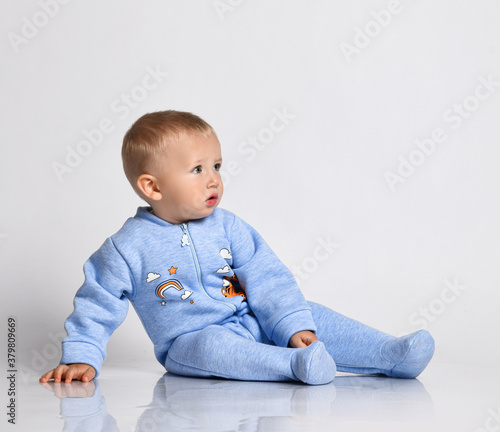 Blond baby boy toddler in blue cotton jumpsuit with rainbow and tiger sits on the floor leaning on his arm