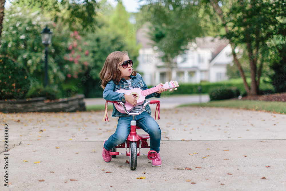 Cute young girl sitting on a tricycle while rocking out with her toy guitar Cute young girl sitting on a tricycle while rocking out with her toy guitar