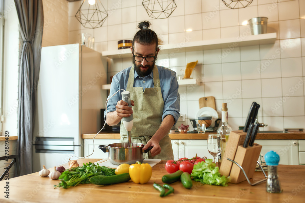 Young man, chef cook using hand blender while preparing Italian meal in the kitchen Young man, chef cook using hand blender while preparing Italian meal in the kitchen