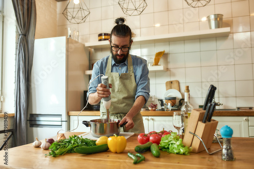 Young man, chef cook using hand blender while preparing Italian meal in the kitchen