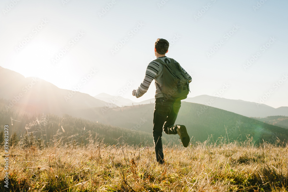 A young tourist man with a backpack run on top and enjoying the sunset. Back view. Hipster walks in the autumn grass in the mountains on vacation. Holiday trip concept. World Tourism Day. A young tourist man with a backpack run on top and enjoying the sunset. Back view. Hipster walks in the autumn grass in the mountains on vacation. Holiday trip concept. World Tourism Day.