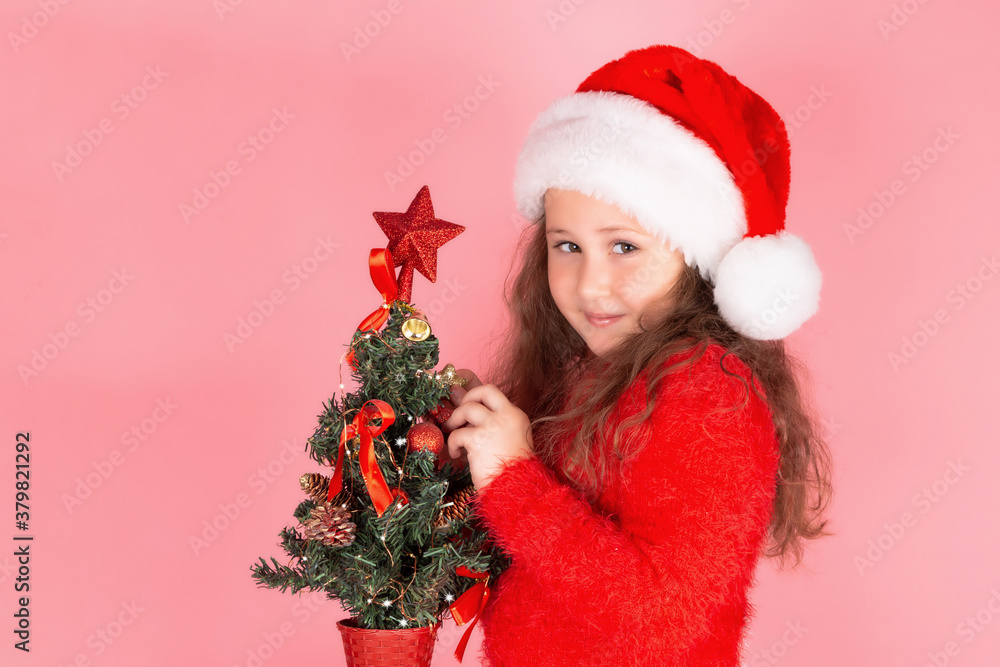 A little girl in a Santa hat is getting ready for the new year and Christmas. A child decorates the Christmas tree with Christmas toys. The concept of a happy childhood. New year 2021. Copy space. 