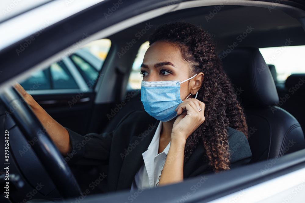 Young business woman fixing and adjusting her medical mask while sitiing in the car behind the steering wheel. Business trips during pandemic, new normal and coronavirus travel safety concept. Young business woman fixing and adjusting her medical mask while sitiing in the car behind the steering wheel. Business trips during pandemic, new normal and coronavirus travel safety concept.
