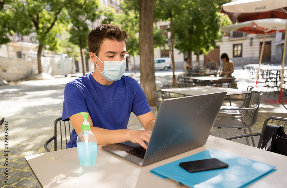 Young man with protective face mask and hand sanitizer outdoors working remotely on computer Young man with protective face mask and hand sanitizer outdoors working remotely on computer