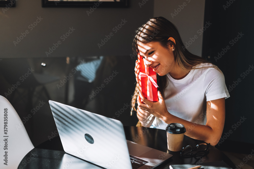 Happy young woman opening gift in front of laptop during video call or chat, celebrating birthday online. Concept of distance relations, celebrations and lifestyle. Happy young woman opening gift in front of laptop during video call or chat, celebrating birthday online. Concept of distance relations, celebrations and lifestyle.