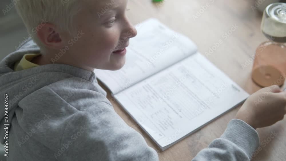 The boy does lessons at home at the table for a while. The child writes with a pen in a notebook and looks at the hourglass. The boy does lessons at home at the table for a while. The child writes with a pen in a notebook and looks at the hourglass.