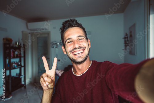 Happy young handsome millennial taking a selfie smiling at the camera in the living room at home.