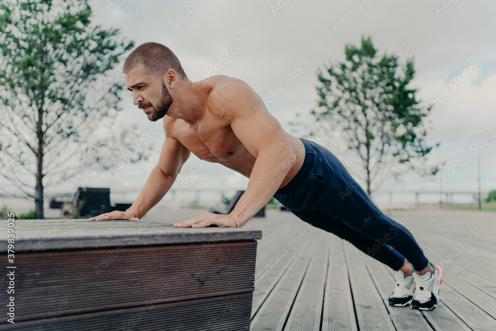 Obraz premium Confident muscled sportsman in sport wear doing push up outdoors, works on biceps and trains determination. Strong topless athletic man exercises in open air. People, motivation, training concept