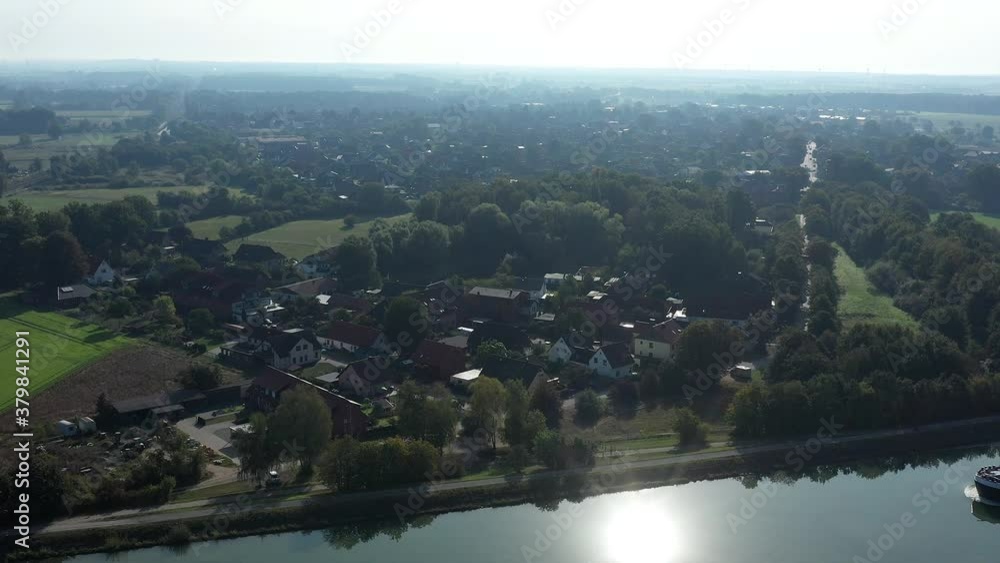 Aerial view of a German village next to a canal on which a cargo ship passes and in which the sun is reflected glittering, with detached houses Aerial view of a German village next to a canal on which a cargo ship passes and in which the sun is reflected glittering, with detached houses