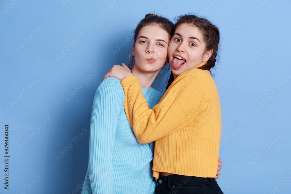 Two positive Caucasian young girlfriends hugging and having fun while posing isolated over blue background, ladies making grimace and showing tongue, wearing casual attires. Two positive Caucasian young girlfriends hugging and having fun while posing isolated over blue background, ladies making grimace and showing tongue, wearing casual attires.