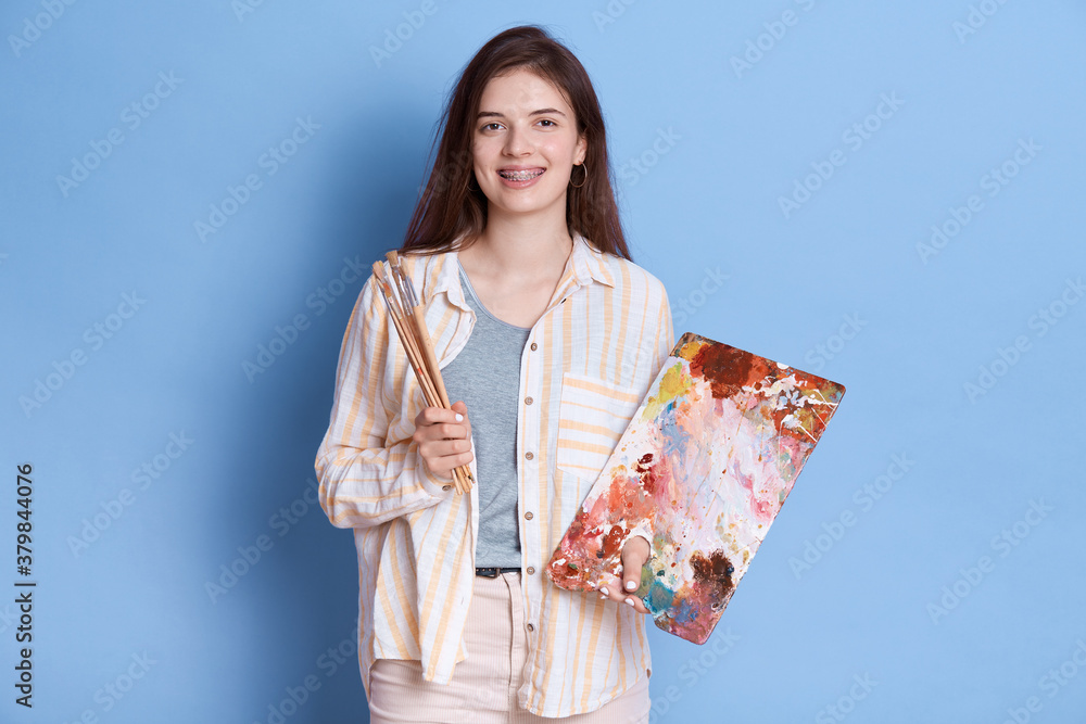 Attractive young Caucasian female artist with painting accessories, holding brushes, mixing colors on palette, lady wearing white shirt and trousers. Attractive young Caucasian female artist with painting accessories, holding brushes, mixing colors on palette, lady wearing white shirt and trousers.