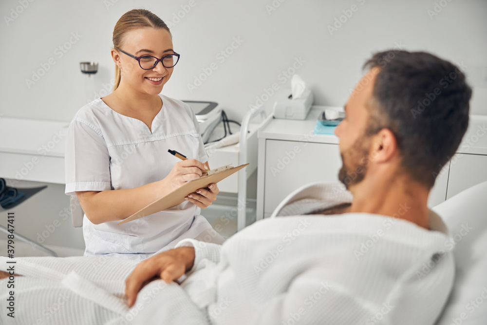 Cheerful beautician talking with male patient and taking notes Cheerful beautician talking with male patient and taking notes
