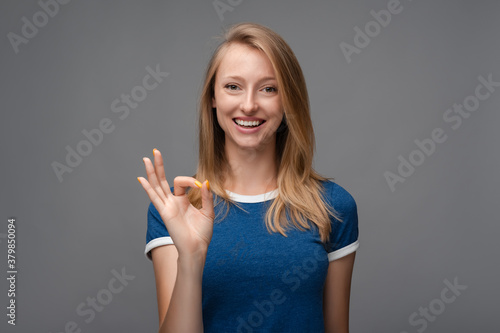 Positive Young blonde woman laughing and showing OK gesture. Studio shot, gray background. Human emotions concept
