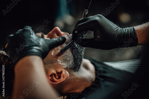 Barber in black gloves maintains hygiene when cutting a beard of his client with sharp razor in shop.