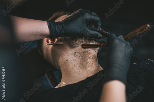 Guy sitting and relaxing in chair while he is shaving by skillful hairdresser with black gloves in cool barbershop.